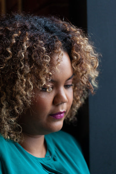 A woman with curly, highlighted hair and wearing a teal top looks down thoughtfully by a dark window, soft natural light illuminating her face.