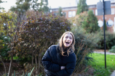 A young woman in a black jacket stands outdoors, laughing joyfully. She is surrounded by greenery and trees, with a blurred building in the background on a bright day.