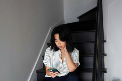 A person with shoulder-length dark hair sits on a staircase, looking down at a smartphone in their hand. They wear a white and black striped shirt and appear thoughtful or focused.