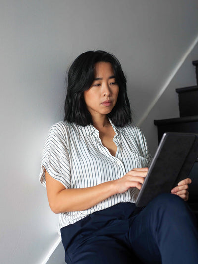 A woman with shoulder-length dark hair sits indoors near stairs, wearing a white and black striped blouse and dark pants, using a tablet and looking at the screen with a focused expression.