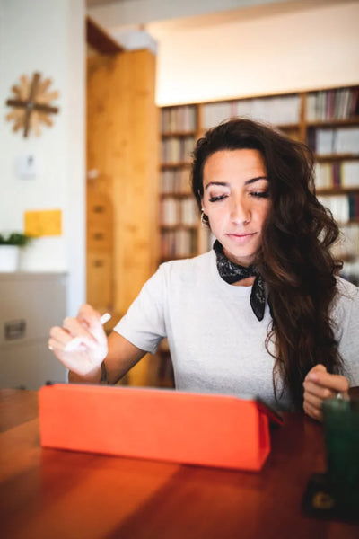 A woman with long dark hair, wearing a white t-shirt and a black bandana, sits at a wooden table using a tablet with an orange case. Bookshelves and a clock are visible in the background.